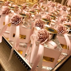 May include: A collection of square glass perfume bottles with gold-toned caps, each adorned with a dusty pink fabric rose and a matching satin ribbon. The bottles feature a textured pink label with a gold rectangular accent on the front surface.