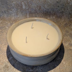 May include: A round, light-coloured ceramic bowl filled with smooth, off-white wax. The candle features three upright white wicks positioned in a triangular formation within the centre of the wax surface. The bowl has a thick, textured rim.