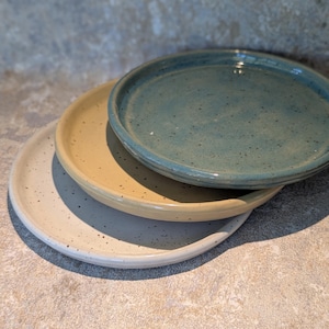 May include: A stack of three round ceramic plates with a speckled finish. The top plate is a deep teal blue, the middle plate is a warm mustard yellow, and the bottom plate is a light cream colour. Each plate features a raised rim and a smooth glaze.