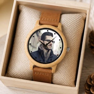 Pu&ograve; includere: Un orologio da polso con cassa in legno chiaro e cinturino in pelle marrone. Il quadrante presenta un ritratto fotografico di una persona con occhiali e giacca scura, con indici neri e lancette nere che indicano l'ora.