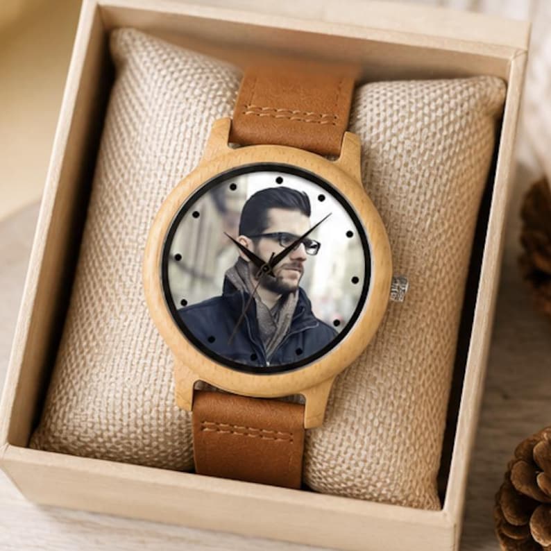 Pu&ograve; includere: Un orologio da polso con cassa in legno chiaro e cinturino in pelle marrone. Il quadrante presenta un ritratto fotografico di una persona con occhiali e giacca scura, con indici neri e lancette nere che indicano l'ora.