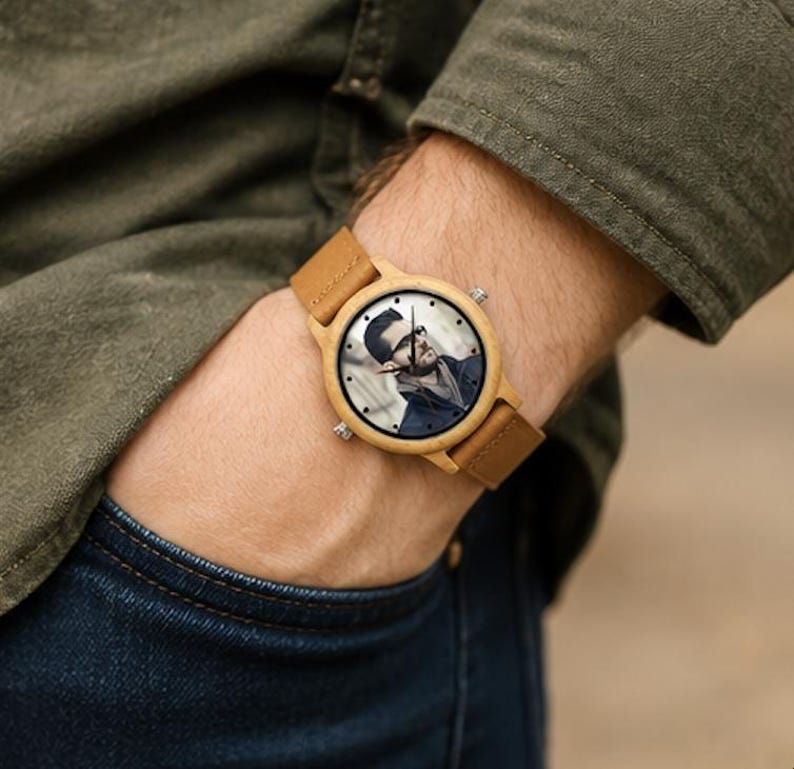 Pu&ograve; includere: Un orologio da polso con cassa in legno marrone chiaro e cinturino in pelle beige abbinato. Il quadrante dell'orologio presenta un ritratto fotografico personalizzato in bianco e nero di una persona con occhiali e lancette nere.