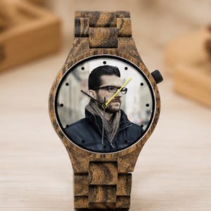 May include: A wristwatch with a dark brown wood grain case and matching link band. The watch face features a photographic portrait of a person wearing glasses and a dark jacket. It has black hour and minute hands and a bright yellow second hand.