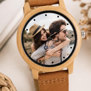 Pu&ograve; includere: Un orologio da polso con cassa in legno chiaro e cinturino in pelle marrone. Il quadrante dell'orologio presenta una stampa fotografica personalizzata di una persona con un cappello di paglia che ne abbraccia un'altra, con indici neri.