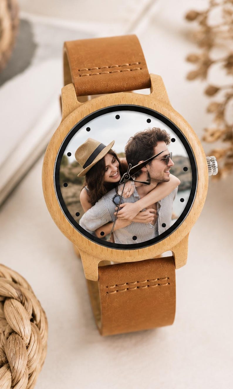 Pu&ograve; includere: Un orologio da polso con cassa in legno chiaro e cinturino in pelle marrone. Il quadrante dell'orologio presenta una stampa fotografica personalizzata di una persona con un cappello di paglia che ne abbraccia un'altra, con indici neri.
