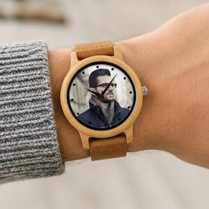 Pu&ograve; includere: Un orologio da polso con cassa in legno chiaro e cinturino in pelle color cuoio. Il quadrante presenta un ritratto fotografico personalizzato di una persona con capelli scuri, barba e occhiali, che indossa una giacca scura e una sciarpa.