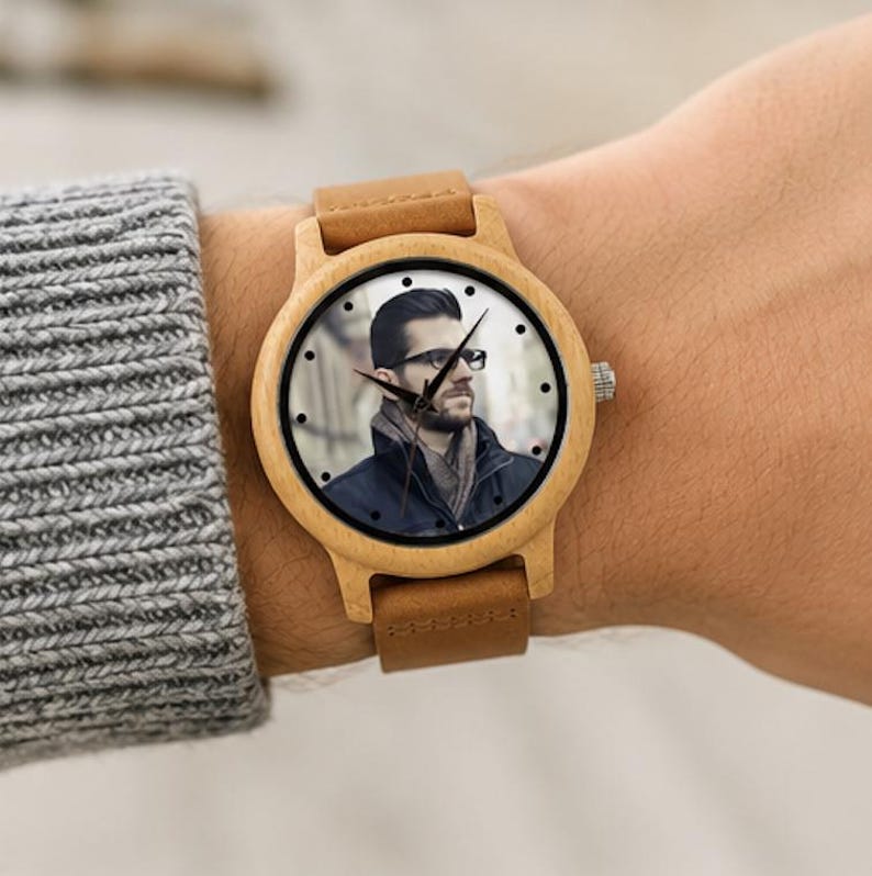 Pu&ograve; includere: Un orologio da polso con cassa in legno chiaro e cinturino in pelle color cuoio. Il quadrante presenta un ritratto fotografico personalizzato di una persona con capelli scuri, barba e occhiali, che indossa una giacca scura e una sciarpa.