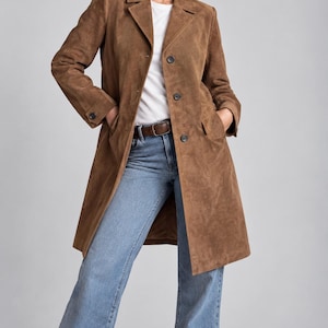 May include: A brown suede coat featuring a notched collar, long sleeves with buttoned cuffs, and a front button closure. The coat is worn over a plain white t-shirt and light blue denim jeans with a brown leather belt at the waist.
