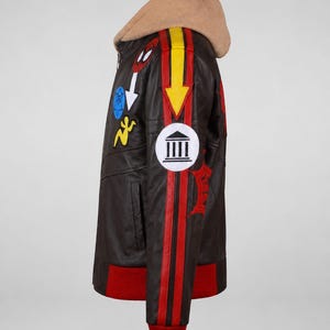 May include: A dark brown leather jacket featuring a tan hood, red ribbed cuffs, and a red hem. The sleeve displays a yellow arrow, a white circular patch with a black temple icon, and various colorful embroidered patches including a red spider mask.