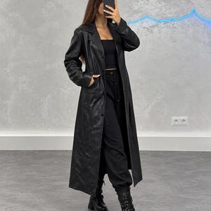 May include: A long black leather coat with a notched collar, long sleeves, and front pockets. The coat is worn over a black strapless top, black drawstring joggers, and black lace-up combat boots with a thick lug sole and side zipper detail.