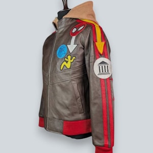 May include: A brown leather jacket featuring a tan hood, red ribbed cuffs and hem, and various colorful patches. The sleeve displays a yellow arrow, red stripes, and a circular patch with a building icon. Additional patches include a blue star and a yellow figure.