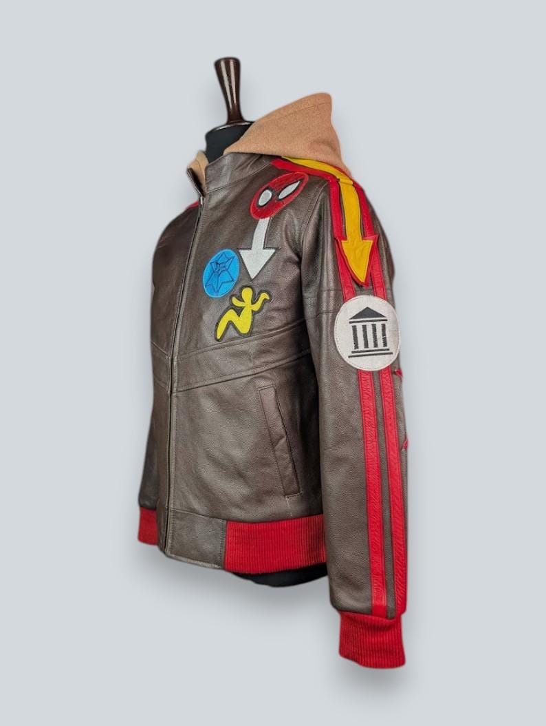 May include: A brown leather jacket featuring a tan hood, red ribbed cuffs and hem, and various colorful patches. The sleeve displays a yellow arrow, red stripes, and a circular patch with a building icon. Additional patches include a blue star and a yellow figure.
