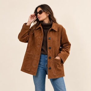 May include: A brown suede jacket featuring a collared neckline, long sleeves, and a button-front closure. The jacket includes two large patch pockets on the front. It is worn over a dark brown turtleneck sweater and blue denim jeans.