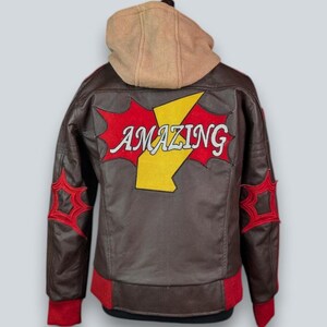 May include: A brown leather jacket featuring a tan hood, red ribbed cuffs, and a red hem. The back displays a large yellow lightning bolt graphic with the word AMAZING in white letters, flanked by red star-shaped patches on each sleeve.