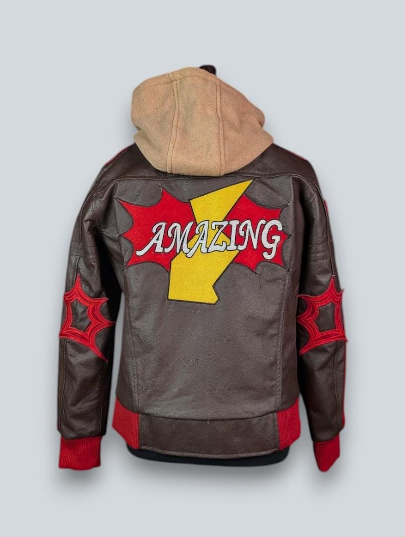 May include: A brown leather jacket featuring a tan hood, red ribbed cuffs, and a red hem. The back displays a large yellow lightning bolt graphic with the word AMAZING in white letters, flanked by red star-shaped patches on each sleeve.