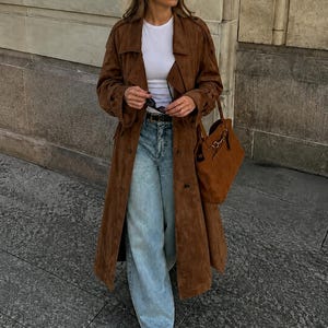 May include: A long brown suede trench coat worn over a white crew neck t-shirt and light blue wide-leg denim jeans. The outfit includes a black belt, a matching brown suede handbag with gold hardware, and black pointed-toe shoes.