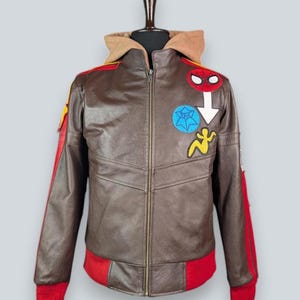 May include: A brown leather jacket featuring a tan hood, a full-length front zipper, and red ribbed cuffs and hem. The chest is decorated with colorful embroidered patches, including a red spider mask, a blue star, and a yellow figure.