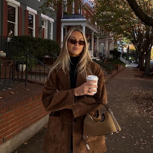 May include: A brown suede blazer with a notched lapel and two large front patch pockets, worn over a black mock neck top. The outfit includes blue denim jeans, dark oval sunglasses, and a small brown handbag with a dark brown leather handle.