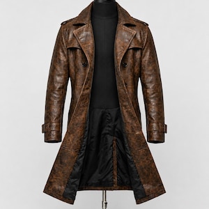 May include: A long, double-breasted trench coat in a distressed brown finish. The coat features wide lapels, shoulder epaulets, and buckled straps at the cuffs. It is displayed on a mannequin with a black interior lining visible at the front opening.