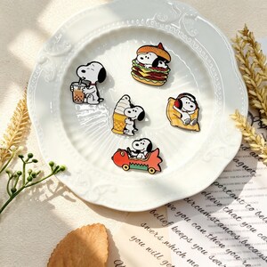 Peut inclure: Un ensemble de cinq broches en émail représentant le personnage de dessin animé Snoopy dans diverses activités : buvant du thé aux perles, assis dans un burger, serrant un cornet de glace, se reposant sur un oreiller jaune et conduisant une voiture rouge.