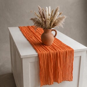 May include: A vibrant orange, crinkled fabric table runner draped over a white surface. A terracotta-colored ceramic pitcher with a braided handle sits on the runner, filled with a bouquet of dried grasses, pampas, and beige floral stems.