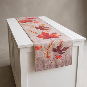 May include: A rectangular table runner with a light brown, textured fabric base. The design features a printed pattern of vibrant red, orange, and dark brown maple leaves interspersed with stalks of golden wheat and thin, dark brown grass blades.