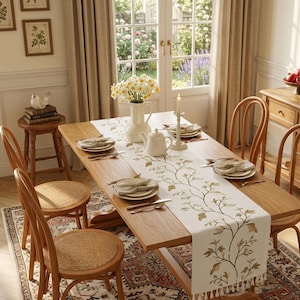 May include: A wooden dining table set with a white table runner featuring an embroidered green and gold vine pattern with fringe ends. The table is arranged with white ceramic plates, a white pitcher, a white vase with yellow flowers, and a candle.