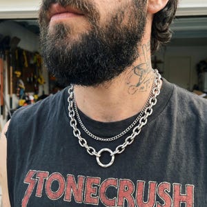 May include: A layered silver-toned necklace set consisting of a thin, fine-link chain and a thicker, heavy-duty oval link chain. The thicker chain features a prominent, circular ring pendant at the center, creating a bold, industrial-style accessory.