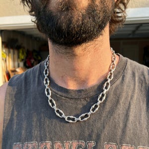Può includere: Una collana a catena color argento con maglie allungate e testurizzate che ricordano l'aspetto del bambù. La collana poggia attorno al collo, mostrando un motivo ripetitivo di barre metalliche segmentate collegate da piccoli anelli lisci.