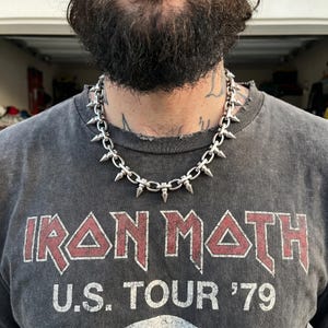 Puede incluir: Un collar de cadena de metal plateado con púas puntiagudas espaciadas uniformemente en cada eslabón. El collar se lleva alrededor del cuello, sobre una camiseta gris oscuro con el texto rojo y blanco IRON MAIDEN U.S. TOUR '79.