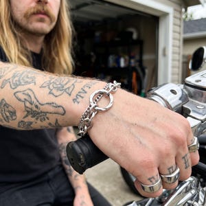 Op de afbeelding: Een zilverkleurige metalen kettingarmband met een opvallende ronde schakel met een gehamerde textuur, verbonden met een reeks kleinere in elkaar grijpende ovale schakels. De armband wordt om de pols gedragen en heeft een gepolijste afwerking.