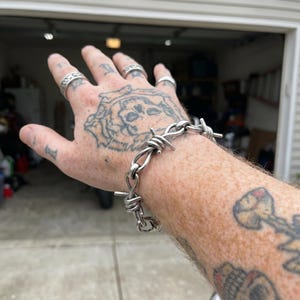 Puede incluir: Una pulsera de cadena de metal plateado diseñada para parecerse al alambre de espino, con eslabones metálicos retorcidos y puntas afiladas que sobresalen. La pulsera se lleva en la muñeca y muestra un acabado metálico pulido y eslabones.