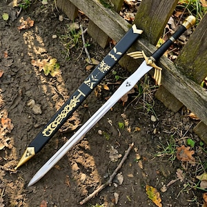 Puede incluir: Una espada de metal con hoja plateada, una guarda dorada con extensiones en forma de alas y un mango negro con pomo dorado. A su lado se encuentra una vaina negra decorada con s&iacute;mbolos dorados, incluido un emblema de trifuerza.