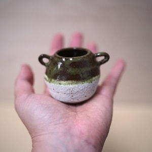 May include: A small, round ceramic pot with two side handles. The upper portion features a glossy, dark brown and olive green glaze, while the lower portion has a textured, matte white finish. The vessel has a wide, open circular rim at the top.