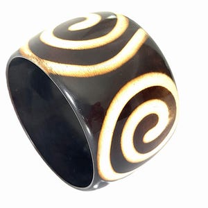 May include: A wide, polished dark brown bangle bracelet featuring a repeating cream-coloured spiral pattern etched into the surface. The bracelet has a smooth, glossy finish and a thick, rounded band construction with a hollow centre.