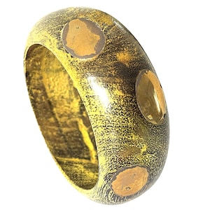 May include: A wide band ring featuring a dark, textured, mottled yellow and black finish. The surface is accented with three smooth, circular, metallic gold-toned inlays spaced evenly around the exterior of the band, creating a contrast.