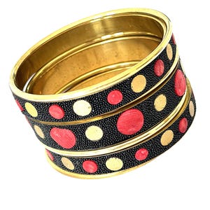 May include: A set of three stacked bangles featuring a gold-toned metal interior and rim. The exterior surface is black with a textured finish, decorated with alternating circular inlays in solid gold-toned metal and vibrant red.