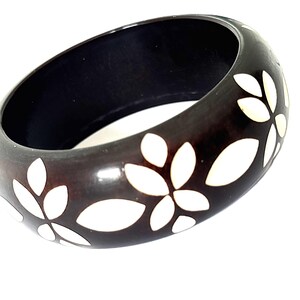 May include: A dark brown, thick bangle bracelet featuring a repeating pattern of white, petal-shaped inlays arranged in floral clusters around the exterior surface. The bracelet has a smooth, polished finish and a wide, rounded band profile.