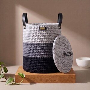 May include: A cylindrical crochet storage basket featuring a colour-blocked design in light grey, dark grey, and black. It includes two black leather handles attached with gold-toned rivets, a small rectangular gold-toned metal tag, and a matching lid.