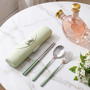 May include: A set of stainless steel cutlery including chopsticks, a spoon, and a fork with sage green handles, accompanied by a matching sage green rectangular storage case featuring a silver bow accent, alongside a glass bottle with a gold cap.