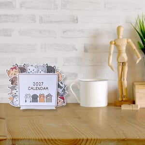 May include: A 2027 desk calendar featuring a rectangular white center with the text 2027 CALENDAR and four cat paws at the bottom. The border is decorated with various illustrated cat faces in different colors, expressions, and accessories.
