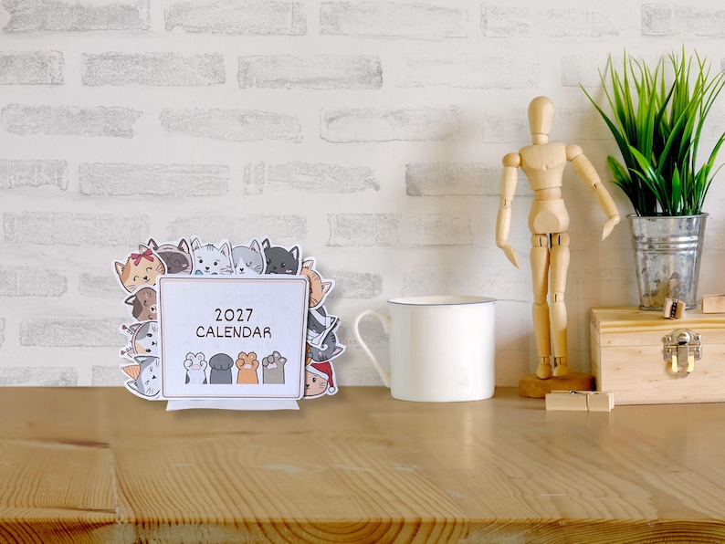 May include: A 2027 desk calendar featuring a rectangular white center with the text 2027 CALENDAR and four cat paws at the bottom. The border is decorated with various illustrated cat faces in different colors, expressions, and accessories.