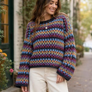 May include: A long-sleeved crochet sweater featuring a multi-colored granny square pattern with horizontal stripes in shades of blue, purple, orange, and green, paired with cream-colored trousers.