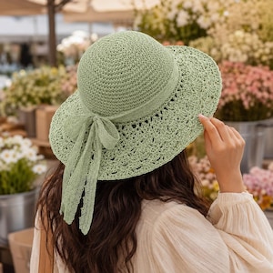 May include: A light sage green sun hat featuring a woven crown and a wide, scalloped lace brim. The hat is accented with a matching sage green ribbon tied into a bow at the back, with two long, delicate lace ribbons hanging down from the bow.