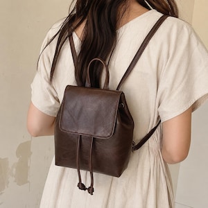 Puede incluir: Una mochila de color marrón oscuro con una textura suave similar al cuero, que presenta una solapa superior con cierre de cordón, un asa superior y dos correas delgadas para los hombros. El bolso tiene una base rectangular estructurada.