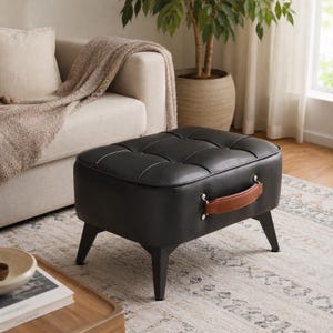 Può includere: Un pouf rettangolare nero caratterizzato da una superficie superiore trapuntata con bottoni. L'oggetto presenta una maniglia color cuoio fissata lateralmente con finiture argentate ed è sostenuto da quattro gambe nere affusolate verso l'esterno.