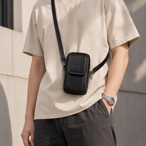 May include: A black vertical crossbody bag with a flap closure and a thin black adjustable shoulder strap. The bag features visible stitching along the edges and is worn over a beige t-shirt and dark grey shorts, with a silver watch on the wrist.