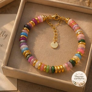 May include: A multi-coloured beaded bracelet featuring alternating smooth round beads in shades of pink, blue, green, and white, interspersed with polished gold-tone metal spacers. It includes a gold-tone lobster clasp and a small round charm.