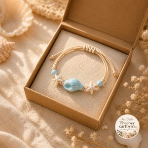 May include: A beige cord bracelet featuring a central light blue ceramic seashell bead flanked by two white starfish charms and two small light blue spherical beads, presented in an open cardboard box with a logo reading Thecozy cartbyiva.