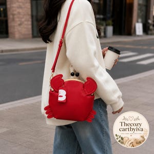 Peut inclure: Un sac bandoulière rouge vif en forme de crabe avec une armature en métal doré, deux perles de fermoir sphériques noires et une bandoulière rouge assortie. Une petite étiquette rectangulaire blanche avec une lettre B rouge est fixée sur le côté.